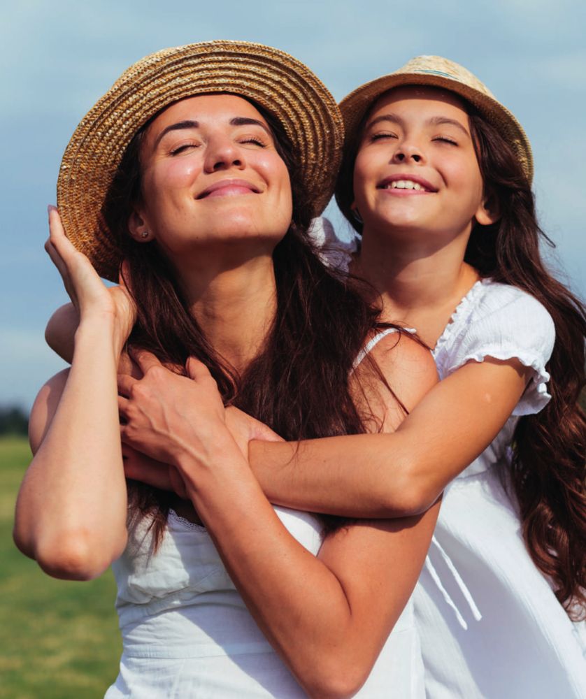 Mãe e filha sorrindo ao sol, abraçadas, usando chapéus de palha